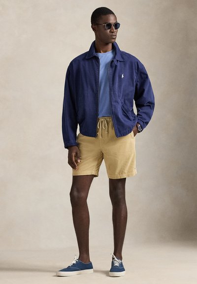 Άνδρας με μπλε σκούρο σακάκι, ανοιχτό μπλε πουκάμισο, μπεζ σορτς, μπλε αθλητικά παπούτσια, γυαλιά ηλίου και ρολόι χειρός, στέκεται μπροστά σε απλό μπεζ φόντο.