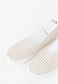Scarpe slip-on a strisce con un motivo bianco e beige, dotate di una suola in gomma e di una tomaia in tessuto morbido. Marchio visibile sul tallone.