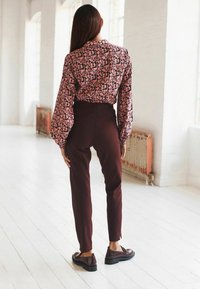 Blouse avec un motif abstrait rose et noir, manches bouffantes ; pantalon ajusté bordeaux foncé ; chaussures plates noires. Intérieur lumineux et minimaliste.
