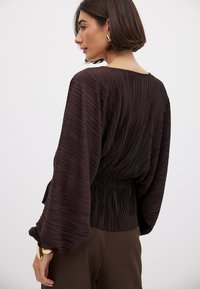 Blusa pieghettata marrone scuro con una vestibilità ampia, vita elastica e maniche larghe. Tessuto morbido con pieghe verticali e minimi dettagli.