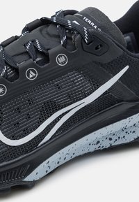 Gros plan d'une chaussure de sport noire et grise avec des lacets à motifs, une bande de logo blanche et une semelle grise tachetée.