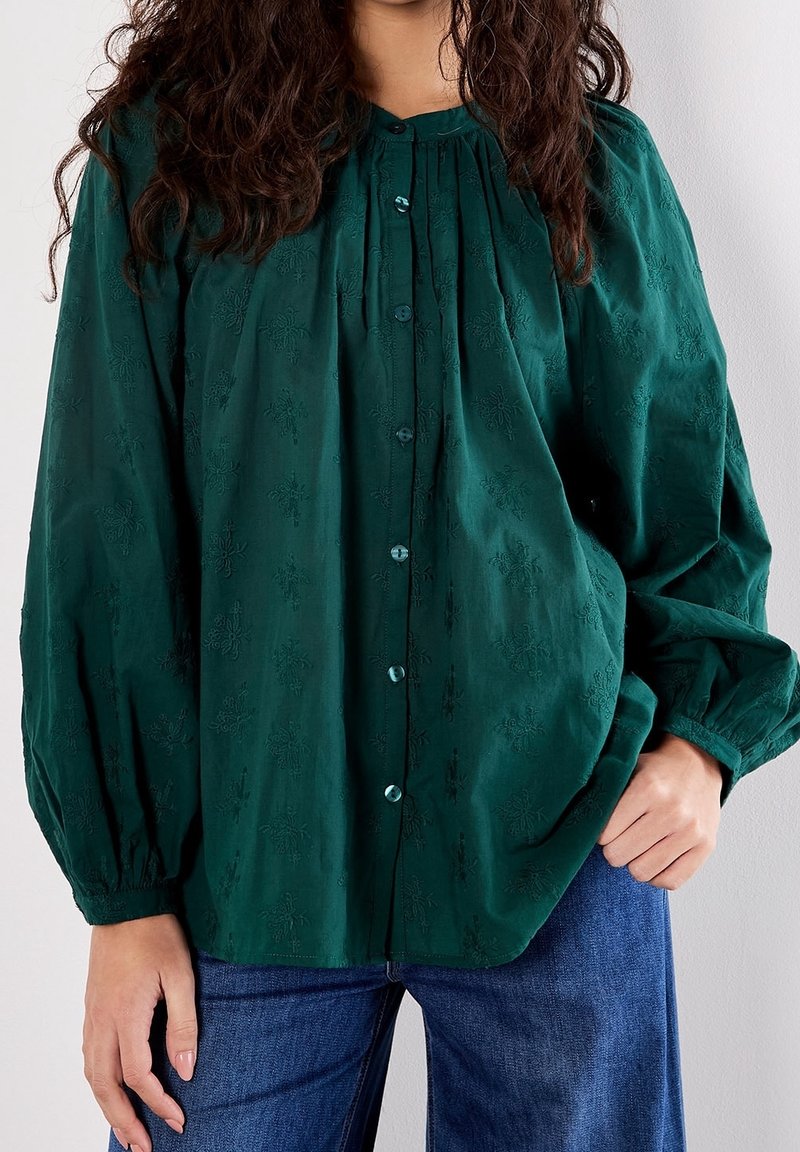 Femme portant un chemisier boutonné vert foncé avec des motifs floraux subtils et des manches bouffantes, associée à un jean bleu, debout devant un fond blanc.