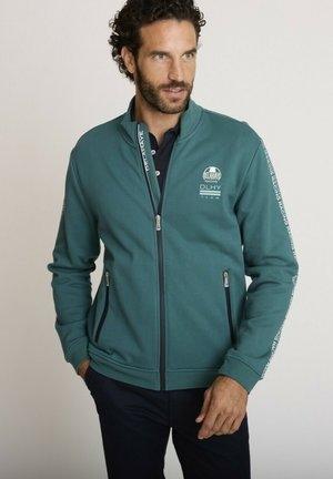 Homme en veste zippée couleur sarcelle avec le logo "Delahaye Racing" et du texte de course sur les manches, regardant légèrement sur le côté avec les mains près des poches.