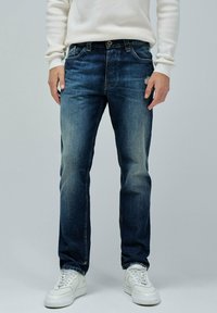 Vaqueros de mezclilla en azul oscuro con un acabado desgastado, diseño de cinco bolsillos y corte recto. Combinados con zapatillas blancas.