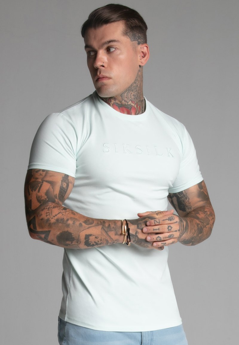 Svjetlucava tanka zelena majica sa kratkim rukavima, koja ima utisnutu "SIKSILK" oznaku na prsima. Glatka tkanina i kroj koji se uklapa.