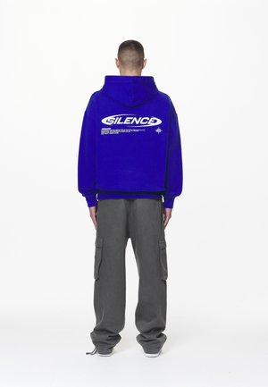 Person trägt einen blauen Kapuzenpullover mit dem Schriftzug „SILENCE“ und einem zusätzlichen kleinen Aufdruck auf dem Rücken, kombiniert mit lockeren grauen Cargohosen und Turnschuhen.