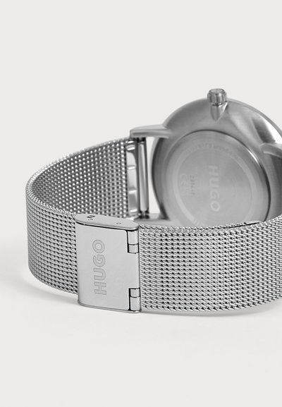 Bracelet de montre en maille métallique argentée avec une boucle gravée "HUGO" et le dos d'un cadran de montre rond en argent visible.