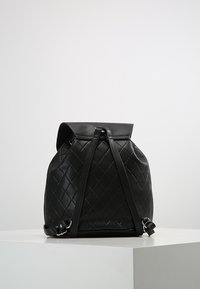 Sac à dos matelassé noir en similicuir, avec rabat supérieur et bretelles ajustables. Texture lisse avec des accents en motif losange.