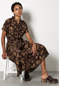Abito camicia con motivo a foglie nero e marrone, con vita cintrata, maniche corte e gonna fluida. Le zeppe in legno marroni completano l'outfit.