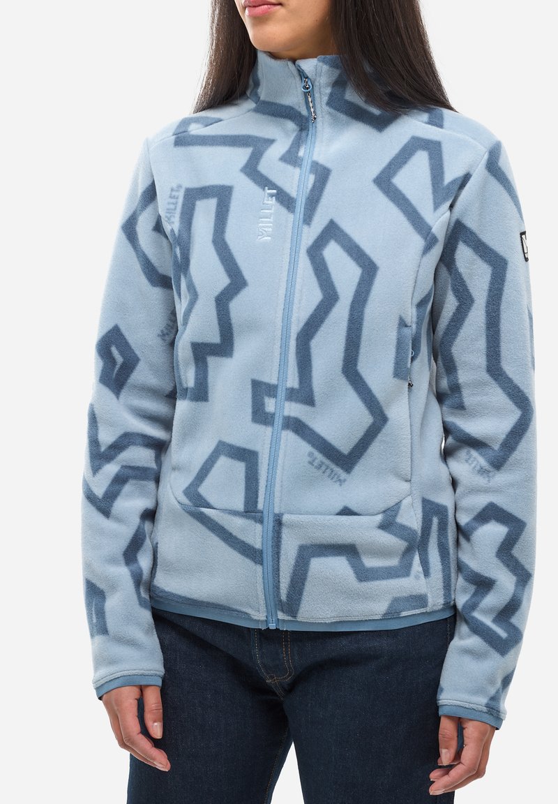 Frau trägt eine hellblaue Fleecejacke mit dunkelblauen abstrakten Zickzackmustern und dem "MILLET"-Logo, kombiniert mit dunkelblauen Jeans.