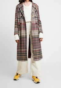 Dlouhý kostkovaný kabát v multikolorovém kostičkovaném vzoru s dvojitým zapínáním, širokými klopami a př oversized střihem. Kombinováno s krémovými kordovými kalhotami a žlutými teniskami.