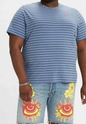 Homme portant un t-shirt à rayures bleues et un short en jean bleu clair décoré d’imprimés d’yeux rouges et jaunes ainsi que de fleurs.