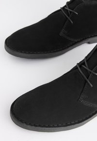 Schwarze Wildleder-Schnürschuhe mit abgerundeten Zehen und aufgenähten Details auf weißem Untergrund.
