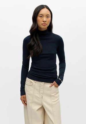 Femme portant un pull à col roulé noir ajusté et un pantalon beige ample avec des coutures visibles, debout avec une main dans la poche sur fond blanc.