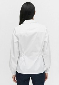 Vit långärmad blus med en figurnära design, strukturerad krage och plisserade detaljer bak. Slät tyg med ribbade manschetter.