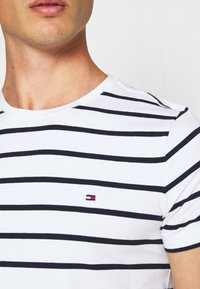 Zbliżenie osoby w białej koszulce z czarnymi poziomymi paskami i małym czerwono-biało-niebieskim logo na piersi.