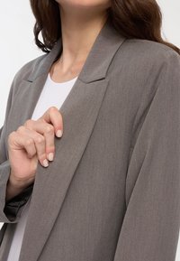 Szara marynarka o dopasowanym kroju, z klapami i miękką teksturą. Model trzyma rękę na klapie marynarki, ukazując wewnętrzną podszewkę.