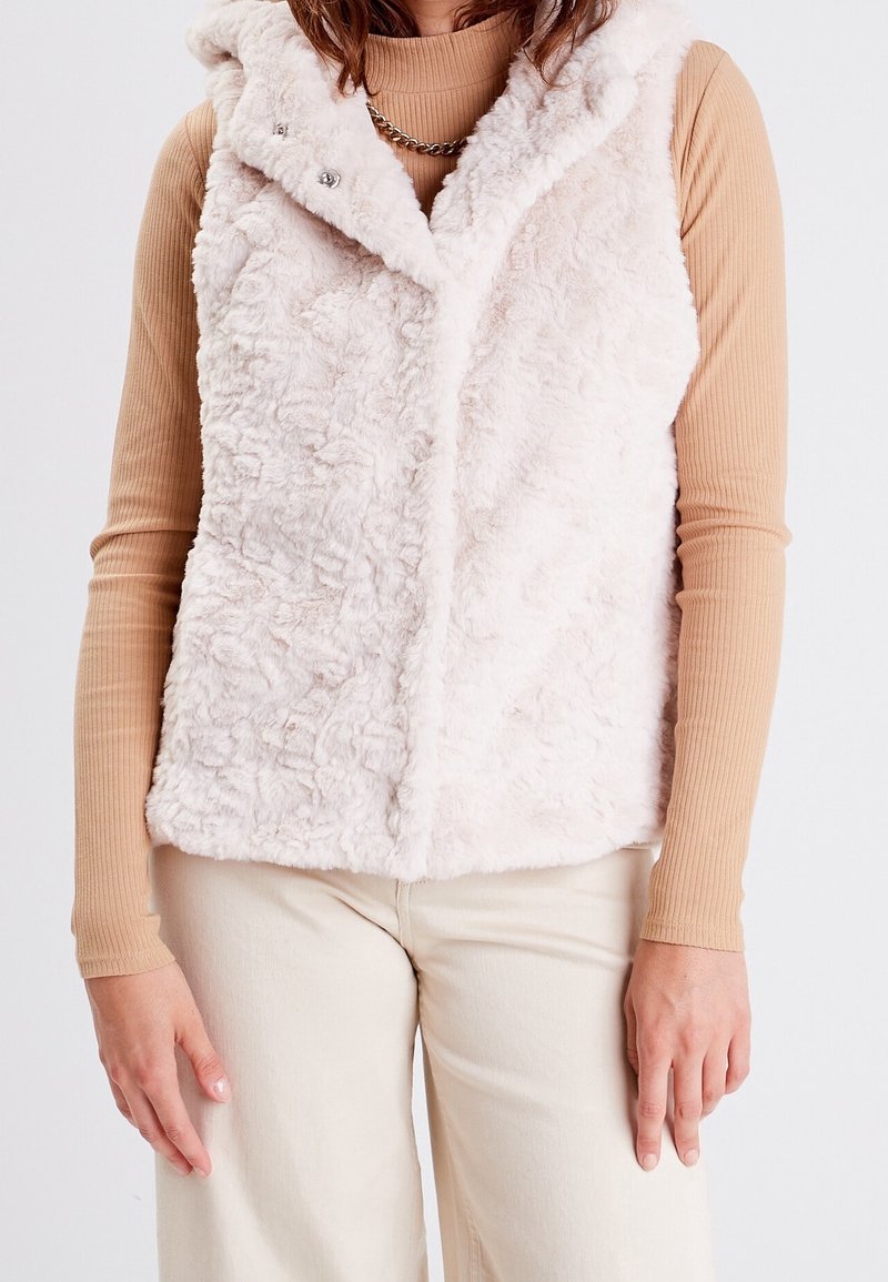 Femme portant une veste blanche sans manches en fourrure par-dessus un haut beige côtelé à manches longues et un pantalon blanc cassé, sur un fond uni.