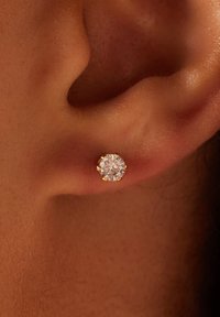 Goldfarbene Stud-Ohrringe mit einem großen, runden, klaren Kristall, der in einem Sechsprong-Design gefasst ist und an einem glatten Ohrläppchen befestigt ist.