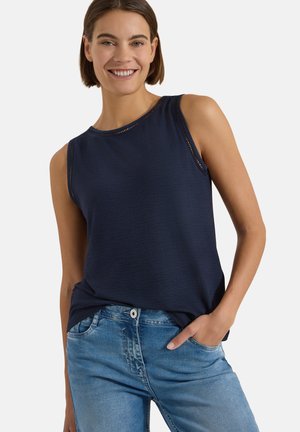 Femme souriante aux cheveux courts et bruns, portant un haut bleu marine sans manches et un jean bleu clair, une main dans la poche, sur un fond uni.