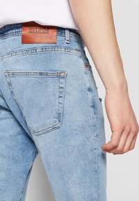 Světle modré džíny z denimu s texturovanou hnědou koženou záplatou s nápisem "2YDNM." Vyznačují se kontrastními švy a zadními kapsami s lehce opotřebovaným vzhledem.