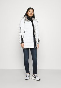 Hvitt puffer-jakke med glidelås foran og hette, med sidelommer og kontrastgrå fôr, parret med mørke jeans og sporty joggesko.