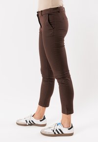 Pantaloni slim-fit marroni con vita a media altezza, tasche laterali e orli risvoltati, abbinati a sneakers bianche e nere. Tessuto dalla superficie liscia.