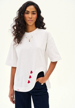 Kobieta z długimi kręconymi włosami, ubrana w białą, oversize'ową koszulkę z asymetrycznym dołem i trzema małymi czerwonymi sercami w pobliżu dołu.