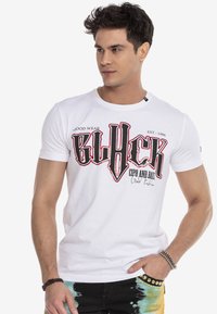Weißes Baumwoll-T-Shirt mit kurzen Ärmeln, das mit einem auffälligen grafischen Textdesign in Schwarz und Rot versehen ist. Das Model trägt eine dunkel gemusterte Hose; Armbänder sind sichtbar.