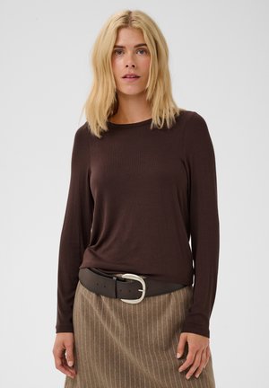 Femme aux cheveux blonds portant un haut à manches longues marron foncé, une large ceinture noire avec une boucle argentée et une jupe beige à fines rayures sur un fond uni.