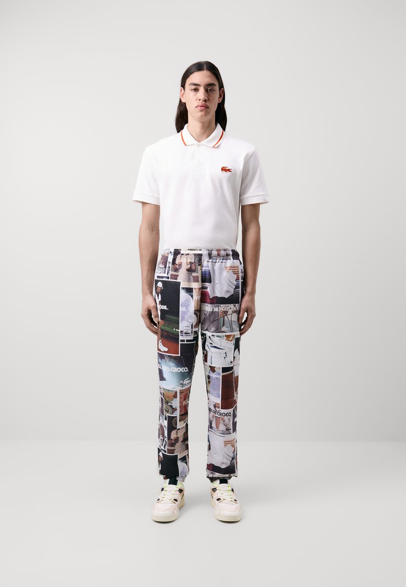 Camisa polo blanca con cuello naranja, combinada con pantalones estampados que presentan impresiones de collage fotográfico. Los zapatos son de color beige claro con acentos negros.