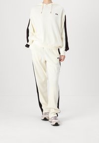 Cremefarbener Hoodie und passende Hose mit schwarzen und roten seitlichen Streifen. Rippstruktur, lockere Passform, kleines Logo-Detail auf der Brust und an der Hose.