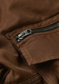 Bruine stof met een gestructureerde afwerking, voorzien van een metalen rits met een trekband. Stiksel in een iets donkerder bruin.