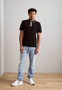 Camiseta polo negra con acentos blancos, de mangas cortas y con un cierre de tres botones, combinada con jeans de color azul claro y zapatillas blancas.