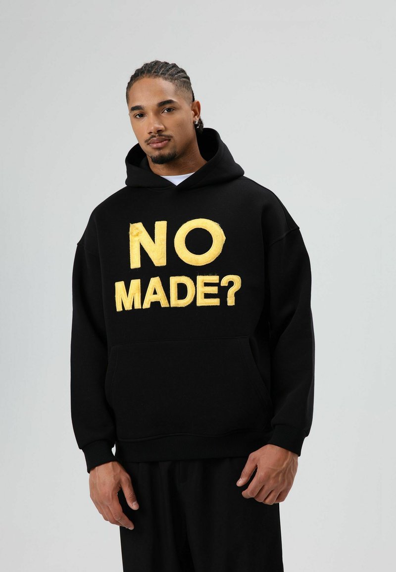 Zwarte hoodie met grote, reliëf gouden tekst "NO MADE?" midden voorop. Heeft een voorkantzak en een ontspannen pasvorm.