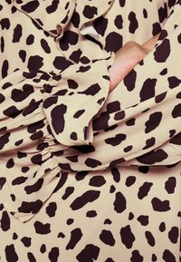 Beige stof met zwarte onregelmatige vlekken, met ruches aan de mouwen en een hand van een persoon die onder de arm is gestoken.