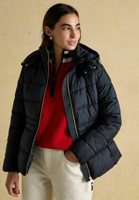 Žena v čiernej prešívanej bunde cez červený sveter s pruhovaným golierom, krémových nohaviciach, s jednou rukou v vrecku, hľadí do strany na pozadí béžovej steny.