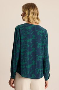 Bluzka z długim rękawem w kolorze granatowym z zielonym wzorem kwiatowym. Miękka tkanina, luźny krój, zaokrąglony dół i mankiety zapinane na guziki. Brak widocznych zapięć.