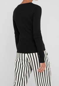 Top nero a maniche lunghe e costine con scollatura tonda, caratterizzato da maniche aderenti e un corpo leggermente svasato. Abbinato a pantaloni a righe bianchi e neri.