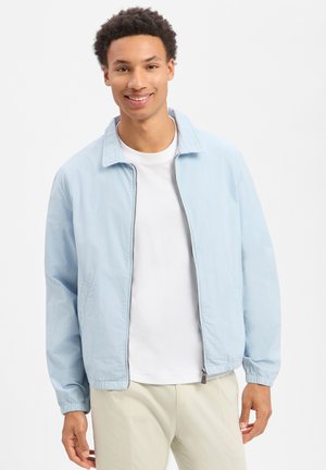 Glimlachende jonge man die een lichtblauw jack met ritssluiting draagt, een wit T-shirt en beige broek, staand tegen een effen achtergrond.