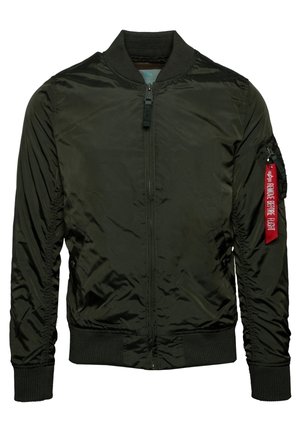 Osoba ubrana w ciemnozieloną kurtkę bomber na białej koszuli i czarnych dżinsach, stojąca z jedną ręką swobodnie opuszczoną wzdłuż ciała.