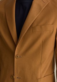 Blazer in cotone marrone con rever a punta, taschino sul petto, due bottoni e finitura testurizzata. Indossato sopra una camicia scura.