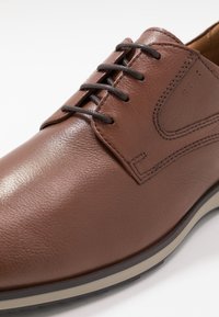 Chaussure en cuir marron à texture lisse, avec des lacets ronds, un design élégant et une semelle en caoutchouc claire contrastante.