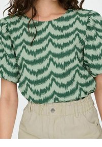 Blouse verte avec un motif en zigzag dans des nuances variées, dotée de manches bouffantes courtes et d'un col rond. Portée sur un pantalon beige clair.