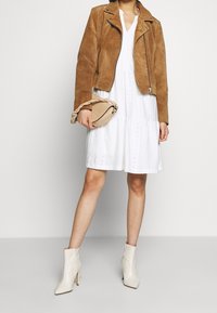 Braune Wildlederjacke über einem weißen Kleid mit Lochstickerei; kombiniert mit einer geflochtenen beigen Tasche und cremefarbenen Knöchelstiefeln mit Reißverschluss.