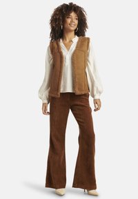 Brown faux suede vest with shearling trim, paired with soft, rust-colored corduroy flared pants. Model wears a white blouse and beige heels.