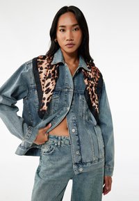 Chaqueta de mezclilla con un acabado azul desgastado, que presenta mangas largas, cierre de botones y una bufanda con estampado de leopardo drapeada alrededor del cuello.