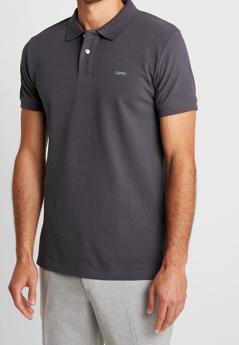Homme portant un polo gris foncé à manches courtes avec deux boutons et un pantalon gris clair, debout devant un fond uni.