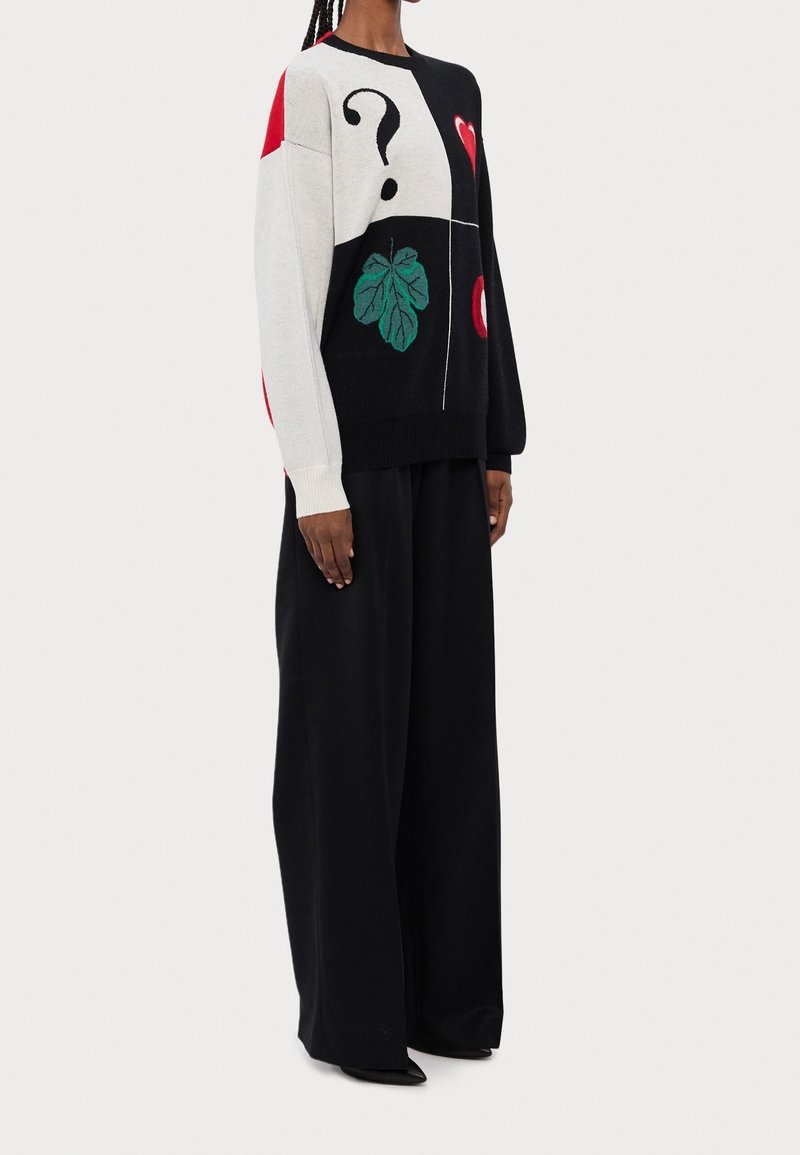 Femme portant un pull noir et blanc avec un point d'interrogation, une feuille verte, des cœurs rouges, associé à un pantalon large noir et des chaussures noires.