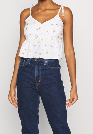 Woman wearing white floral sleeveless crop top with tie straps and high-waisted dark blue jeans against plain background.